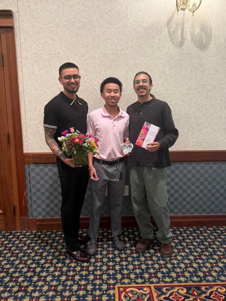 Three men standing indoors, one holding colorful flowers, another holding a red gift box, posing together for a photo